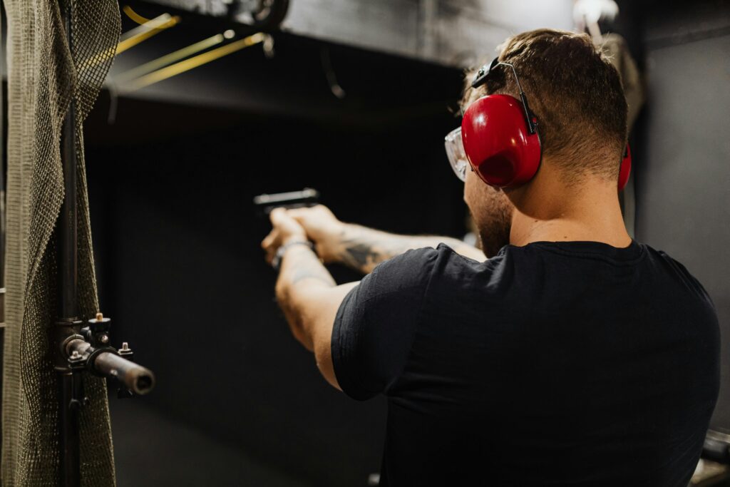 A man practicing competitive shooting at an indoor range, wearing red earmuffs and safety glasses, aiming a handgun.